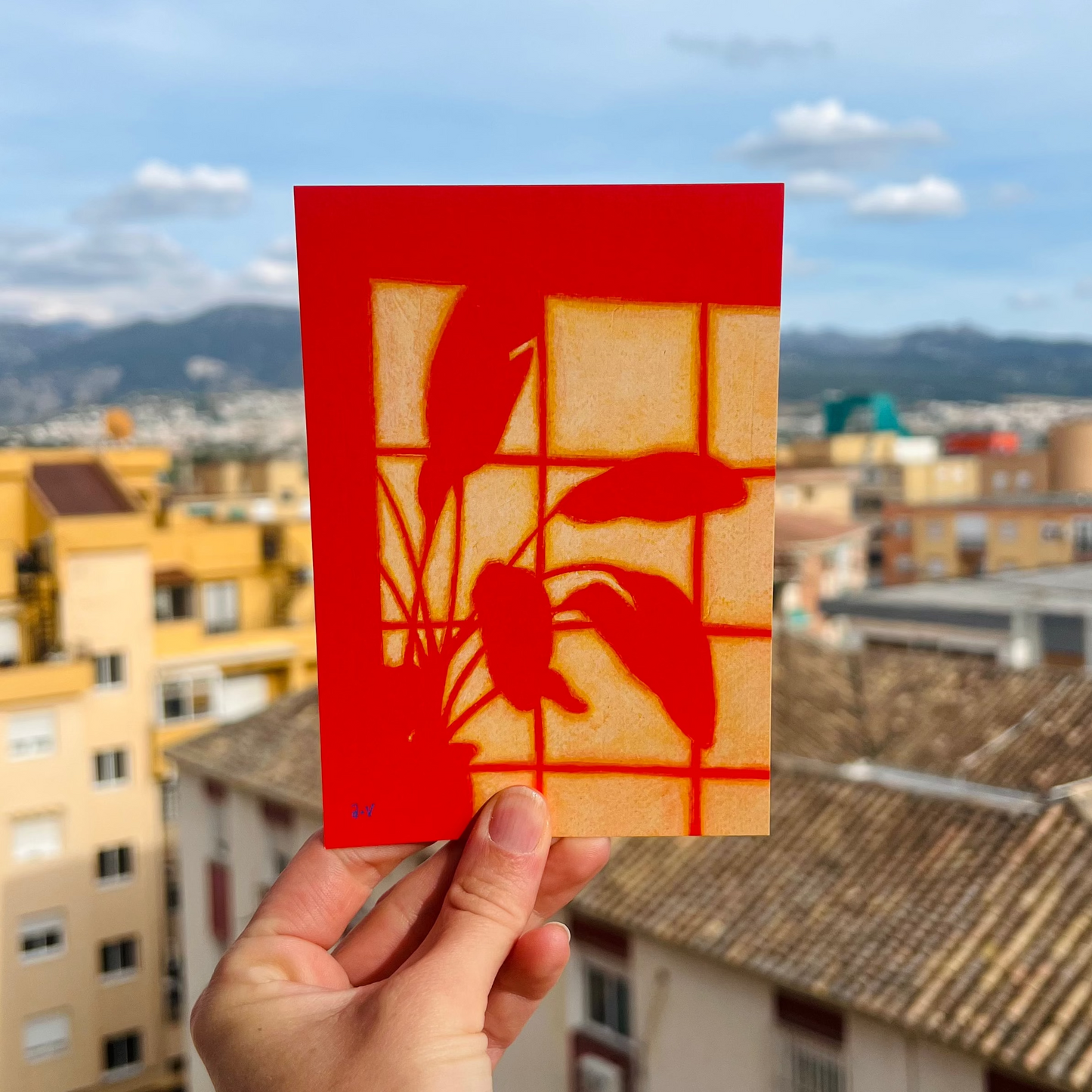 Hand holding a postcard of the shadow of a plant on a wall, against a cityscape background